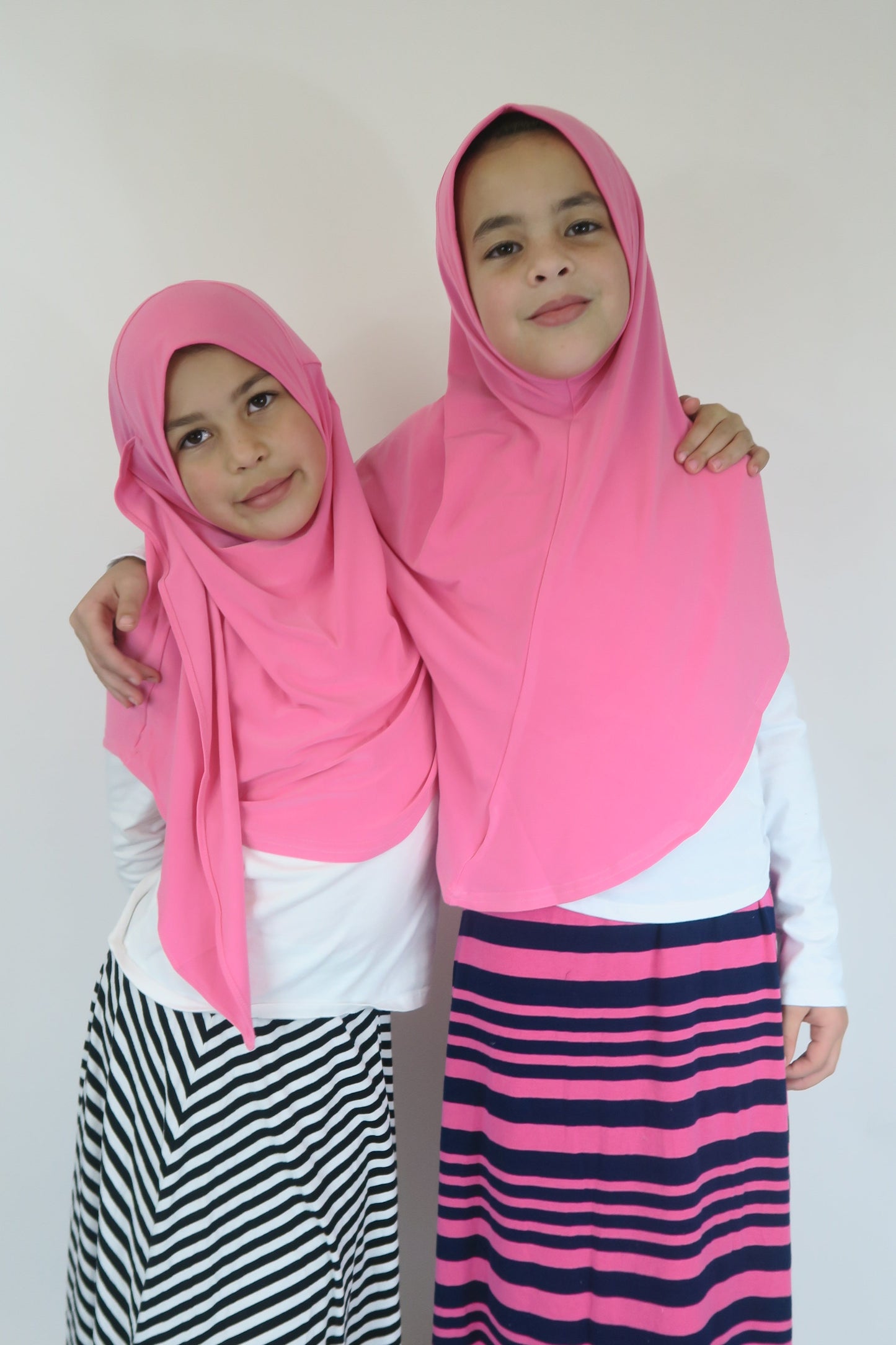 Two people wearing pink hijabs standing side by side against a white background